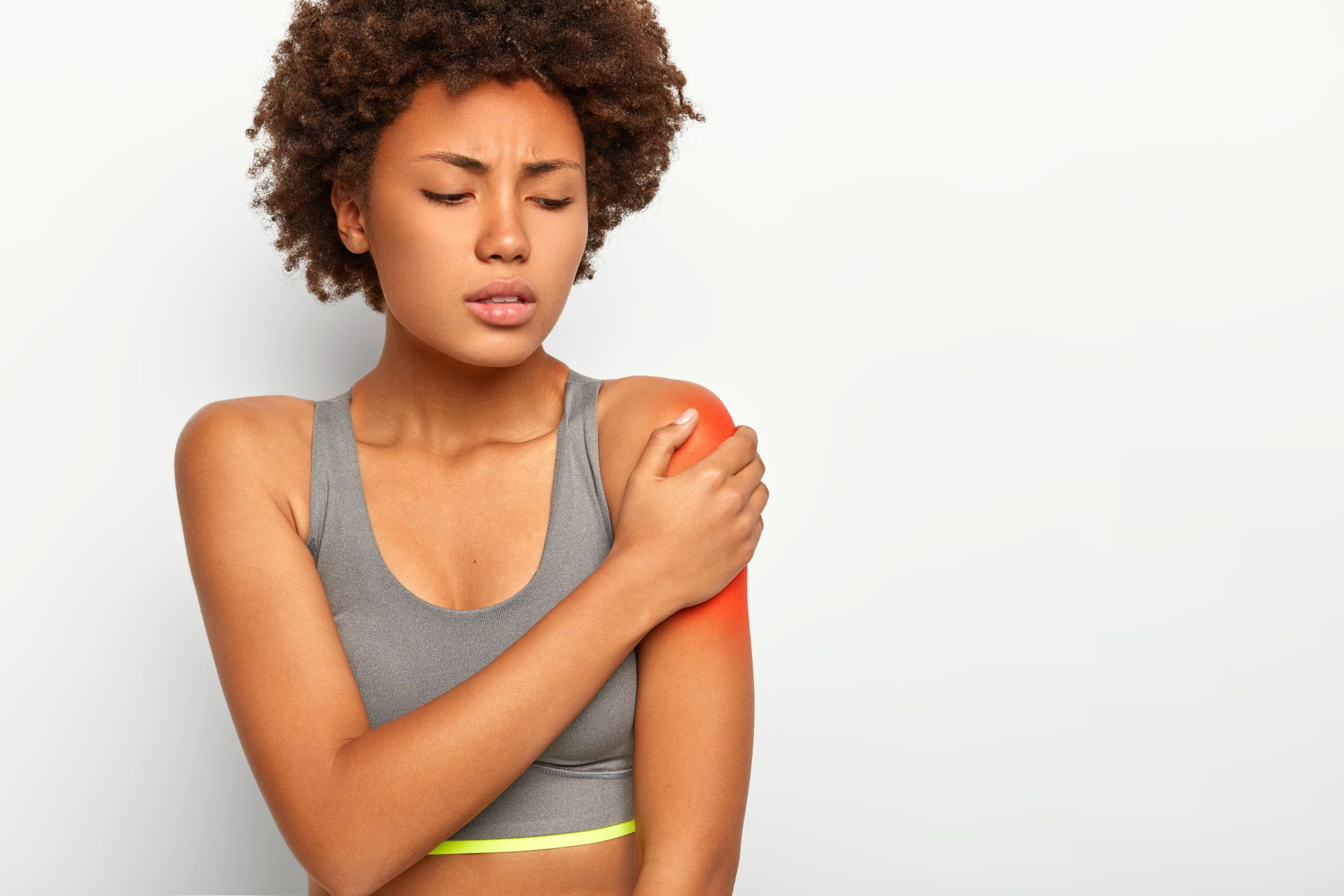 Horizontal shot of dissatisfied Afro woman touches red shoulder, stretched muscles during sport training, has sad expression, wears grey bra, isolated over white background. Health problems.